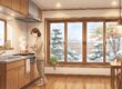 雪が積もる冬の庭を二重窓越しに眺めながら暖かいキッチンで料理をする女性のイラスト。木目調のインテリアと断熱効果のある二重窓が描かれている。